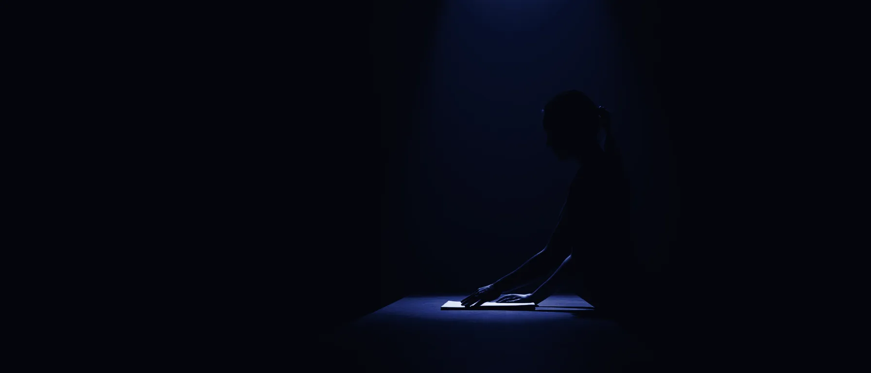 A woman alone at a long table in a dark studio, one beam of blue light overhead