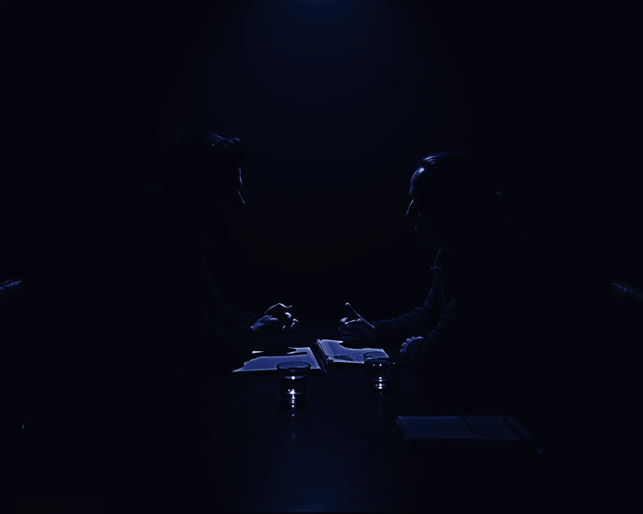 Two people talking across a dark table under a single blue light